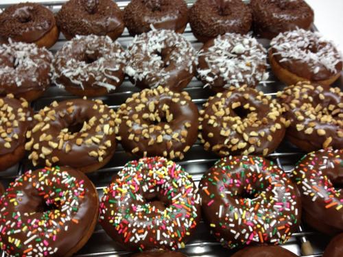 A variety of chocolate-covered donuts topped with sprinkles, coconut, and nuts, arranged on a cooling rack.