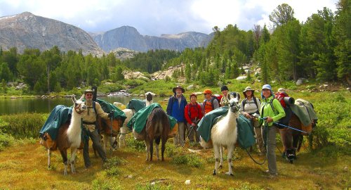 A group of hikers with pack llamas stands by a lake, surrounded by mountains and trees on a cloudy day.