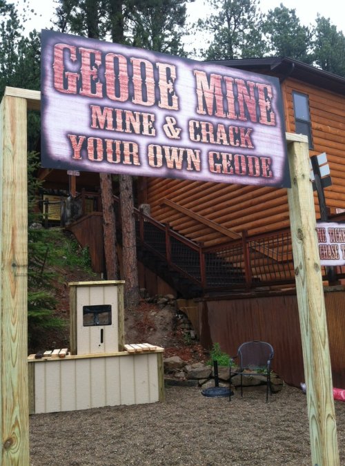 Sign for "Geode Mine" inviting visitors to mine and crack their own geodes, with a rustic wooden building in the background.