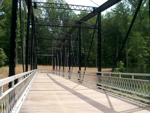 A metal truss bridge with a wooden deck, surrounded by trees and open fields, casting long shadows on the ground.