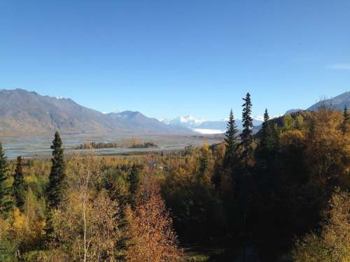 A scenic view of mountains and a valley, with autumn foliage and a clear blue sky. Snow-capped peaks in the distance.