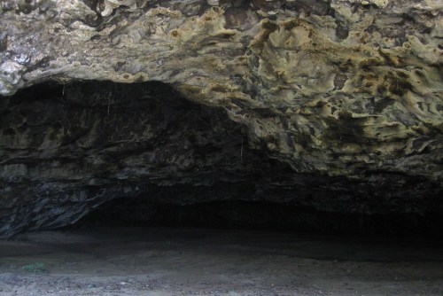 A dark cave entrance with textured rock formations and a sandy floor, surrounded by natural vegetation.