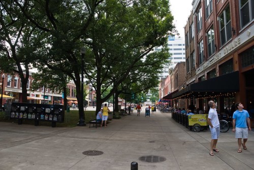 A lively urban street scene with trees, outdoor dining, and people walking in a vibrant area.