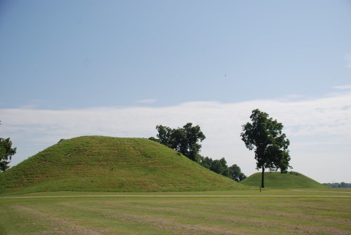 Two large grassy mounds under a clear blue sky, surrounded by trees and open fields.