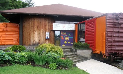 A wooden building with a sign, surrounded by greenery and two colorful train cars parked nearby.