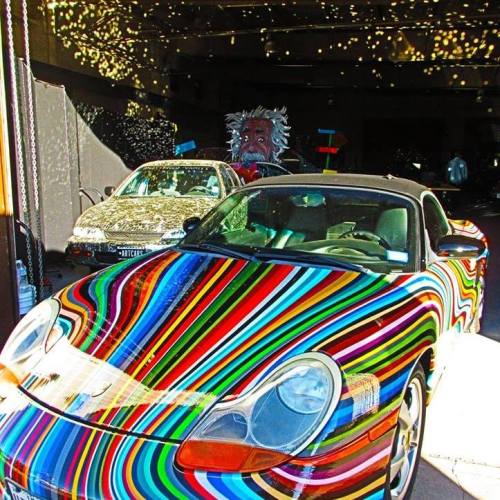 A colorful, striped sports car parked next to a car, with a mural of Einstein in the background.
