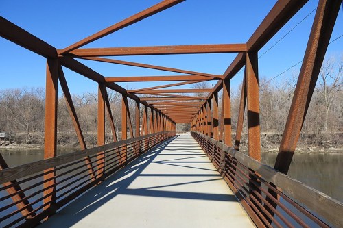 A metal bridge with a rusted frame spans over a river, surrounded by bare trees under a clear blue sky.