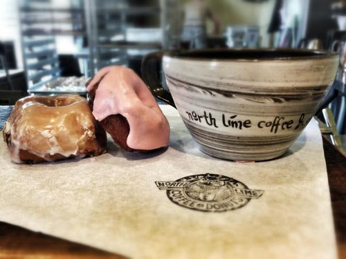 A coffee cup and two donuts, one glazed and one with pink icing, on a paper with a logo from North Lime Coffee & Donuts.