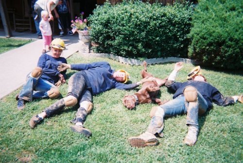 Three people in helmets lie on the grass with a dog, all looking relaxed and playful, surrounded by greenery.
