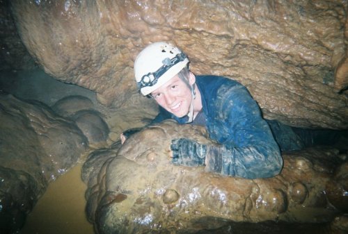 A person in a helmet smiles while crawling through a narrow cave passage, surrounded by rocky formations and water.