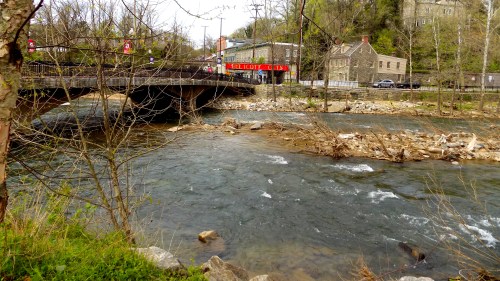 A river flows under a bridge, with trees and rocks along the banks, and a historic building in the background.