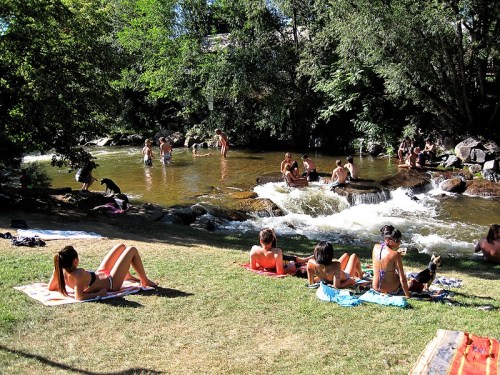 People enjoying a sunny day by a river, some swimming, others sunbathing on towels by the water's edge.
