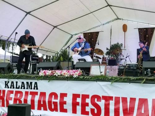 A band performs on stage at the Kalama Heritage Festival, with musicians playing guitar and singing under a tent.