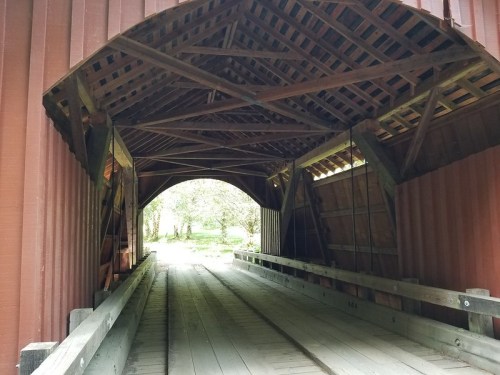 View inside a wooden covered bridge, showcasing the rustic beams and a pathway leading to a bright, green landscape.