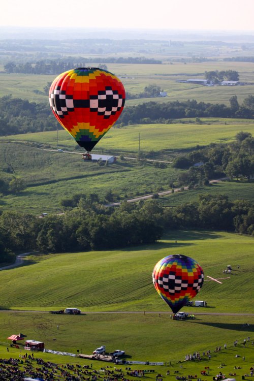 Two colorful hot air balloons float over a green landscape, with spectators gathered below on a sunny day.