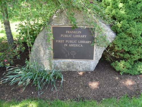 A stone monument with a plaque reading "Franklin Public Library, First Public Library in America," surrounded by greenery.