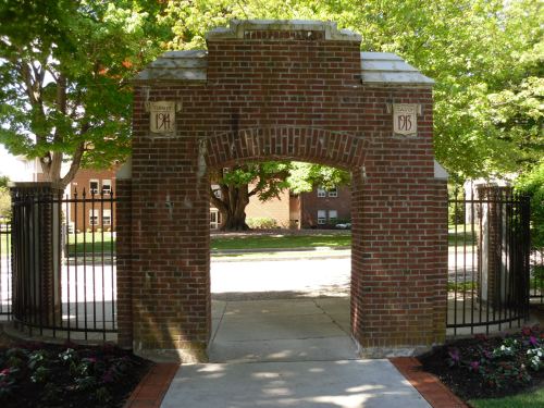 Brick archway with wrought iron gates, leading to a tree-lined pathway and buildings in the background.