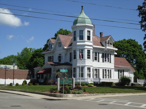 A large, white Victorian house with a green dome, surrounded by trees and a well-maintained lawn, on a sunny day.
