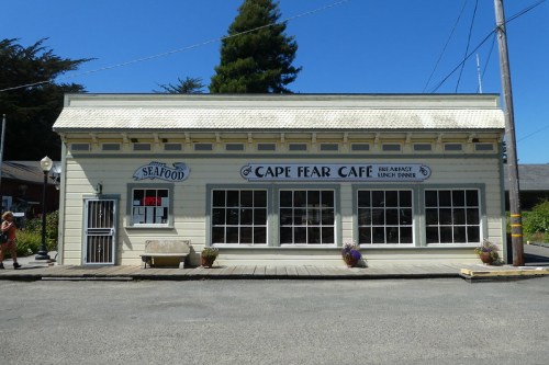 A quaint café building with large windows, featuring signs for "Cape Fear Café" and "Seafood," set on a sunny street.