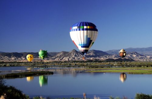Colorful hot air balloons float over a serene lake, reflecting mountains and clear blue skies in the background.