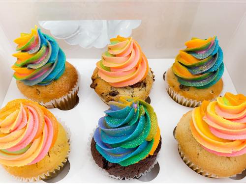 A box of six colorful cupcakes with swirled rainbow frosting, featuring chocolate chip and chocolate bases.
