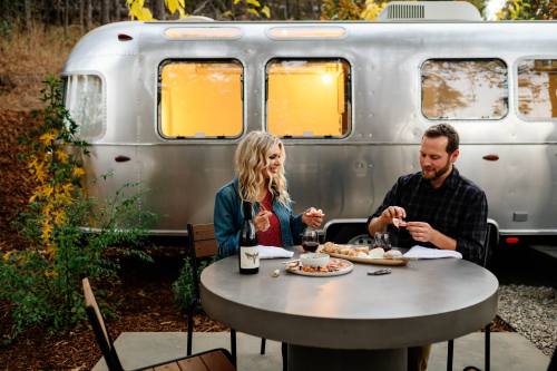 A couple enjoys a meal at a table outside an Airstream trailer, surrounded by nature in autumn.