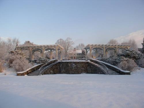 A snow-covered landscape featuring a stone structure with arches and steps, surrounded by trees and a clear sky.