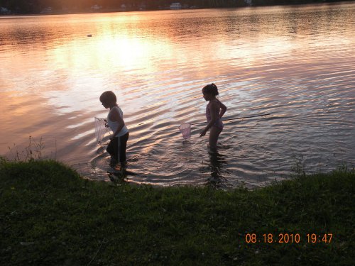 Two children wade in a lake at sunset, one with a net, the other in a swimsuit, surrounded by calm water and reflections.