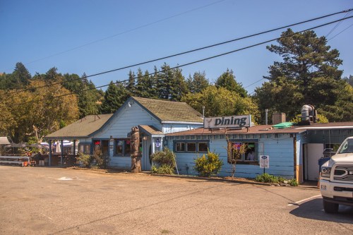 A blue building with a "Dining" sign, surrounded by trees and a gravel parking area on a sunny day.