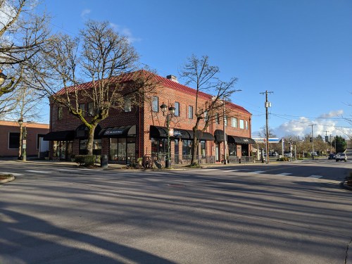 A brick building with a red roof at a street corner, featuring large windows and awnings, under a clear blue sky.