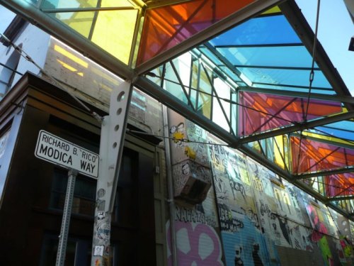 Colorful glass panels overhead illuminate a street sign for Richard B. Rico Modica Way, with vibrant graffiti on the walls.