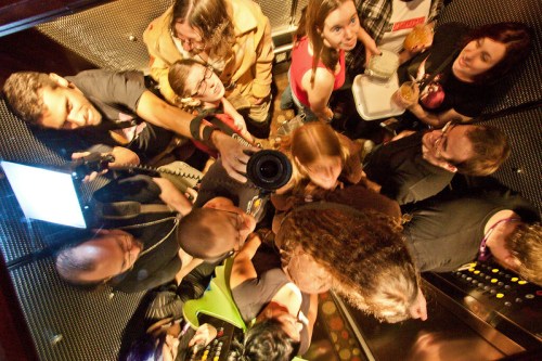 A crowded elevator filled with people taking photos and enjoying snacks, with reflections and vibrant colors.