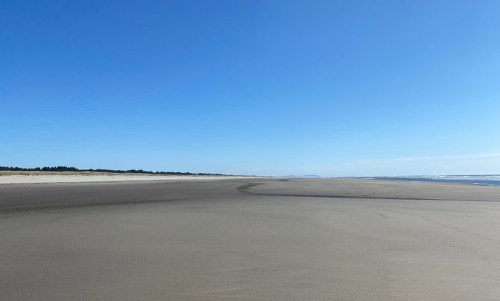 A wide, empty beach under a clear blue sky, with gentle waves lapping at the shore in the distance.
