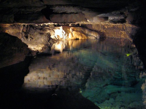 A dimly lit cave with a calm, reflective pool of water and rocky formations surrounding it.