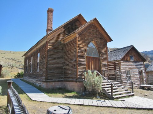 A rustic wooden building with a tall chimney, featuring an arched window and a wooden walkway leading to the entrance.