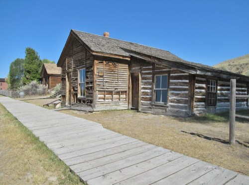 A rustic wooden cabin with a slanted roof, surrounded by grass and a wooden walkway under a clear blue sky.