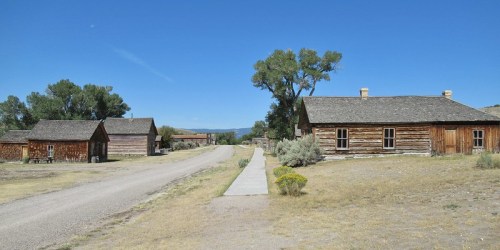 A dirt road lined with rustic wooden buildings and trees under a clear blue sky.