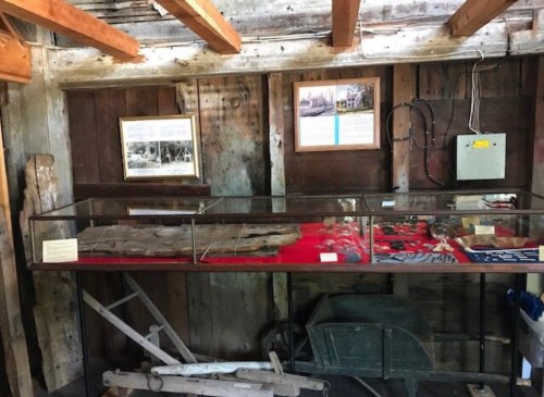 Display case with historical artifacts, wooden tools, and informational plaques in a rustic wooden interior.