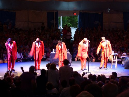 A vocal group in red suits performs on stage in front of a large audience under a tent.