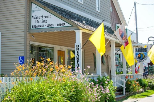 A charming waterfront restaurant with colorful flags and flowers, featuring a sign for breakfast, lunch, and dinner.