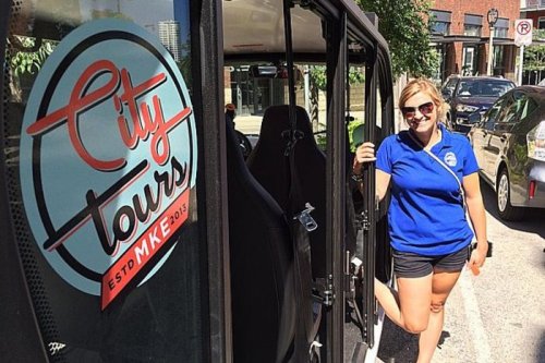 A smiling woman in a blue shirt stands by a city tour bus with a logo that reads "City Tours ESTD MKE 2015."