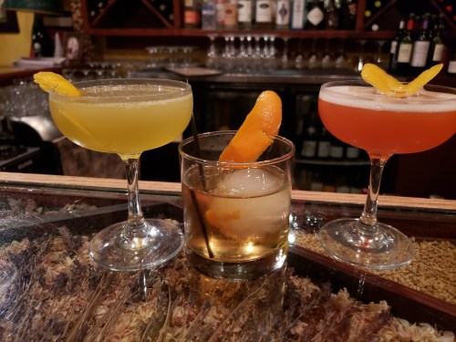 Three colorful cocktails on a bar: a yellow drink, an orange drink with a twist, and a red drink, all garnished with citrus.