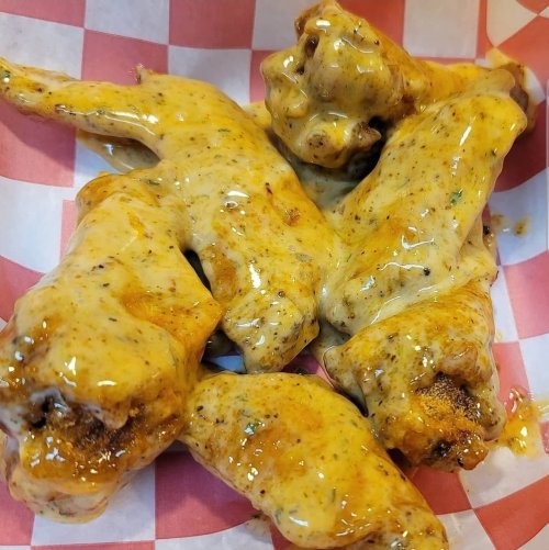 A close-up of several chicken wings coated in a creamy, yellow sauce, served in a red and white checkered paper basket.