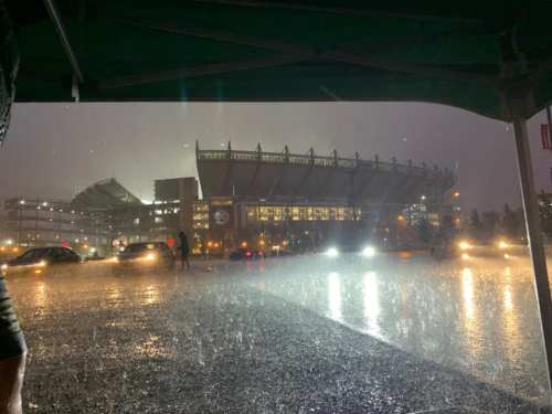 Heavy rain pours down on a stadium, with cars and people visible in the downpour under a dark sky.