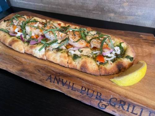 A flatbread topped with shrimp, herbs, and vegetables, served on a wooden board with a lemon wedge.