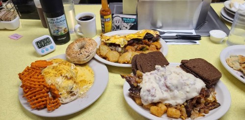 A table with three plates of hearty breakfast food: eggs, bagel, tater tots, and a meat dish with gravy on bread.
