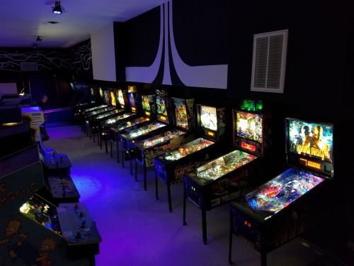 A row of colorful pinball machines illuminated in a dimly lit arcade, featuring various game themes and designs.