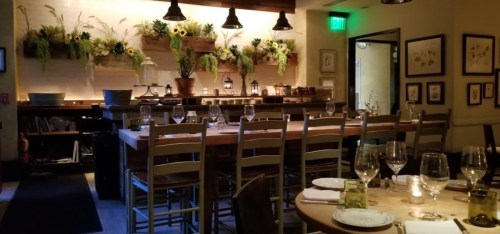 A cozy restaurant interior with a long wooden table, glassware, and a floral wall display, softly lit ambiance.