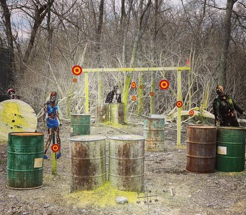 A paintball course with barrels, targets, and figures in camouflage among trees and brush.