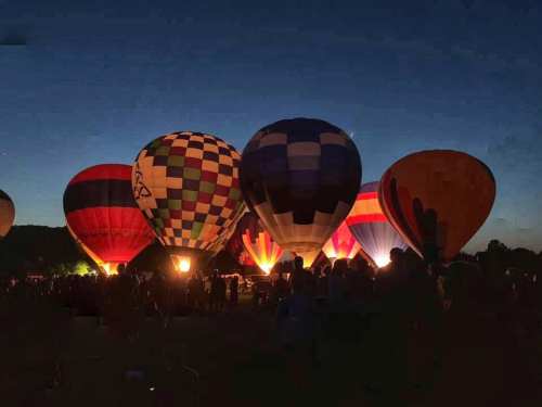 Colorful hot air balloons illuminated against a twilight sky, with a crowd gathered below.